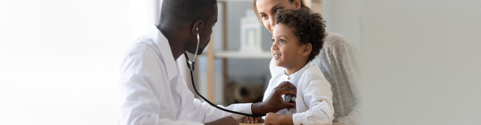 African American male pediatrician with stethoscope listening to lung and heart sound of little boy sitting on mother lap